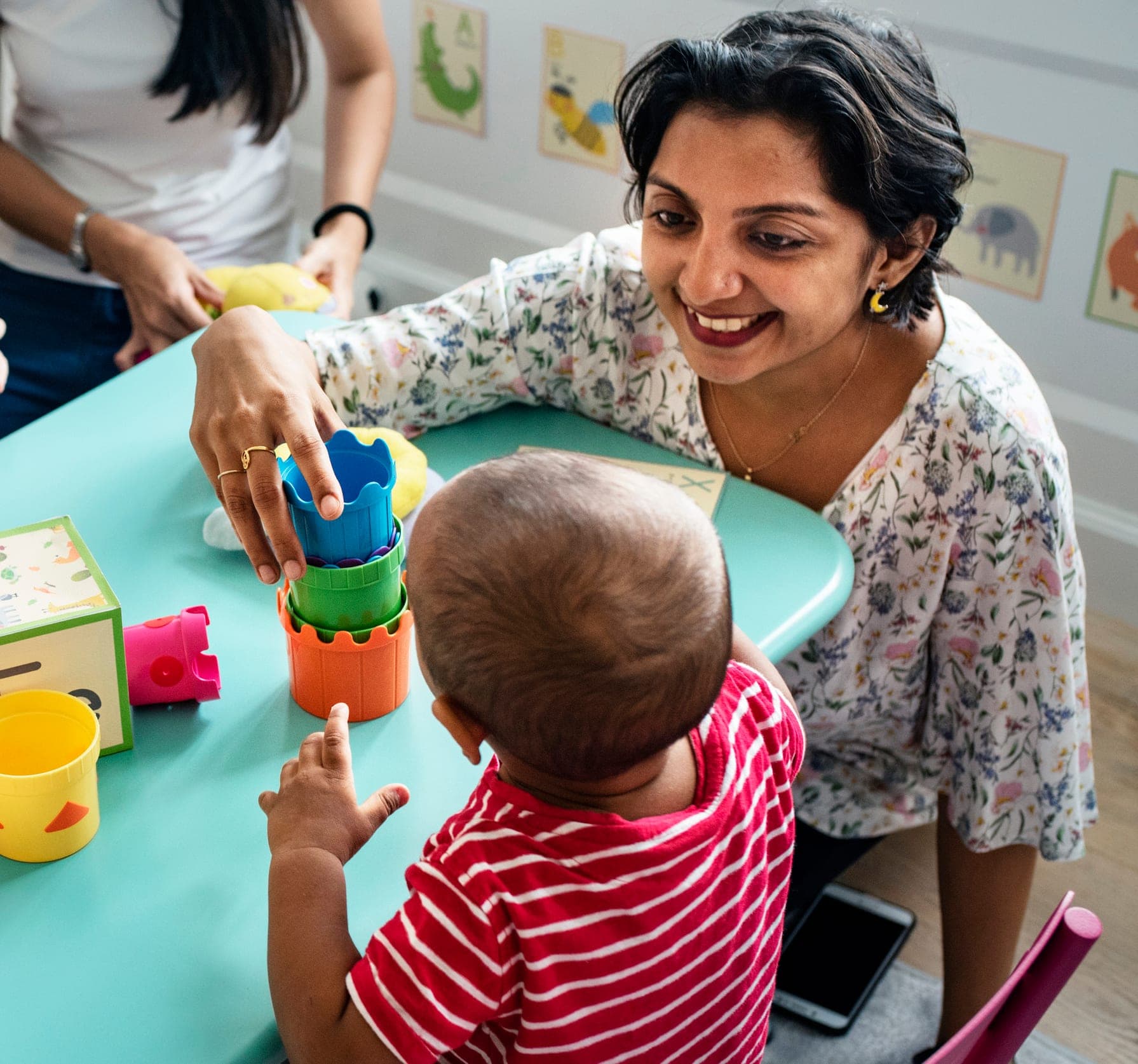 A Little Giants teacher smiling with a baby stacking colorful cups at a classroom table